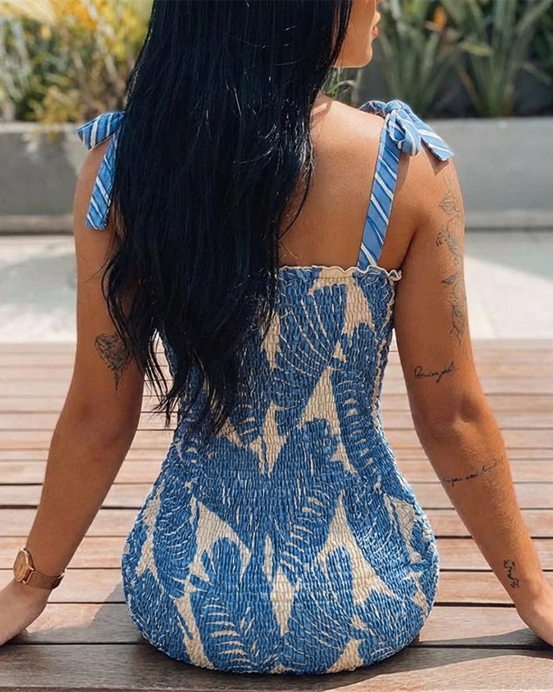 A tropical print mini dress with bow tie shoulders and frills, showing arm tattoos.