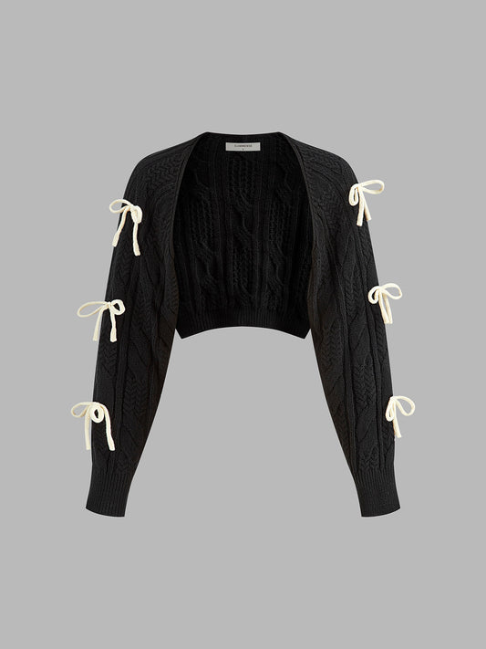 A black cropped cable knit cardigan with open front and three white ribbon bows per sleeve.